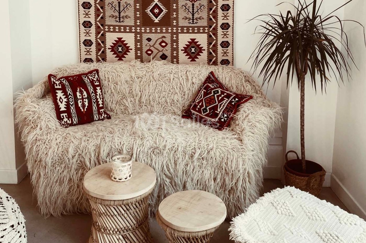 Canapé recouvert de tissu texturé blanc, décoré de coussins rouges à motifs, avec tables en bois et plante en pot.