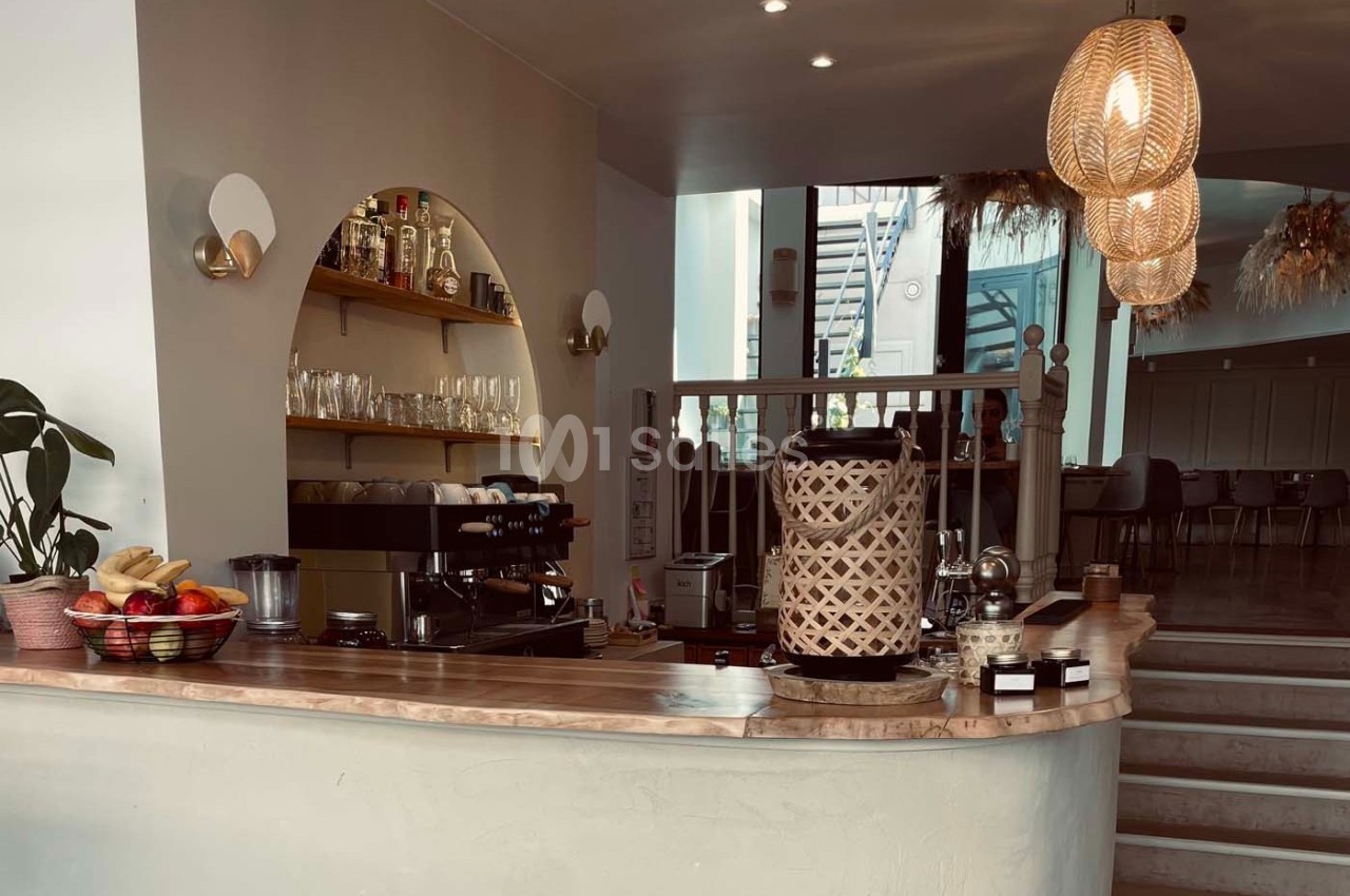 Comptoir de café avec machine à expresso, paniers décoratifs, fruits et étagères contenant des bouteilles et des verres.