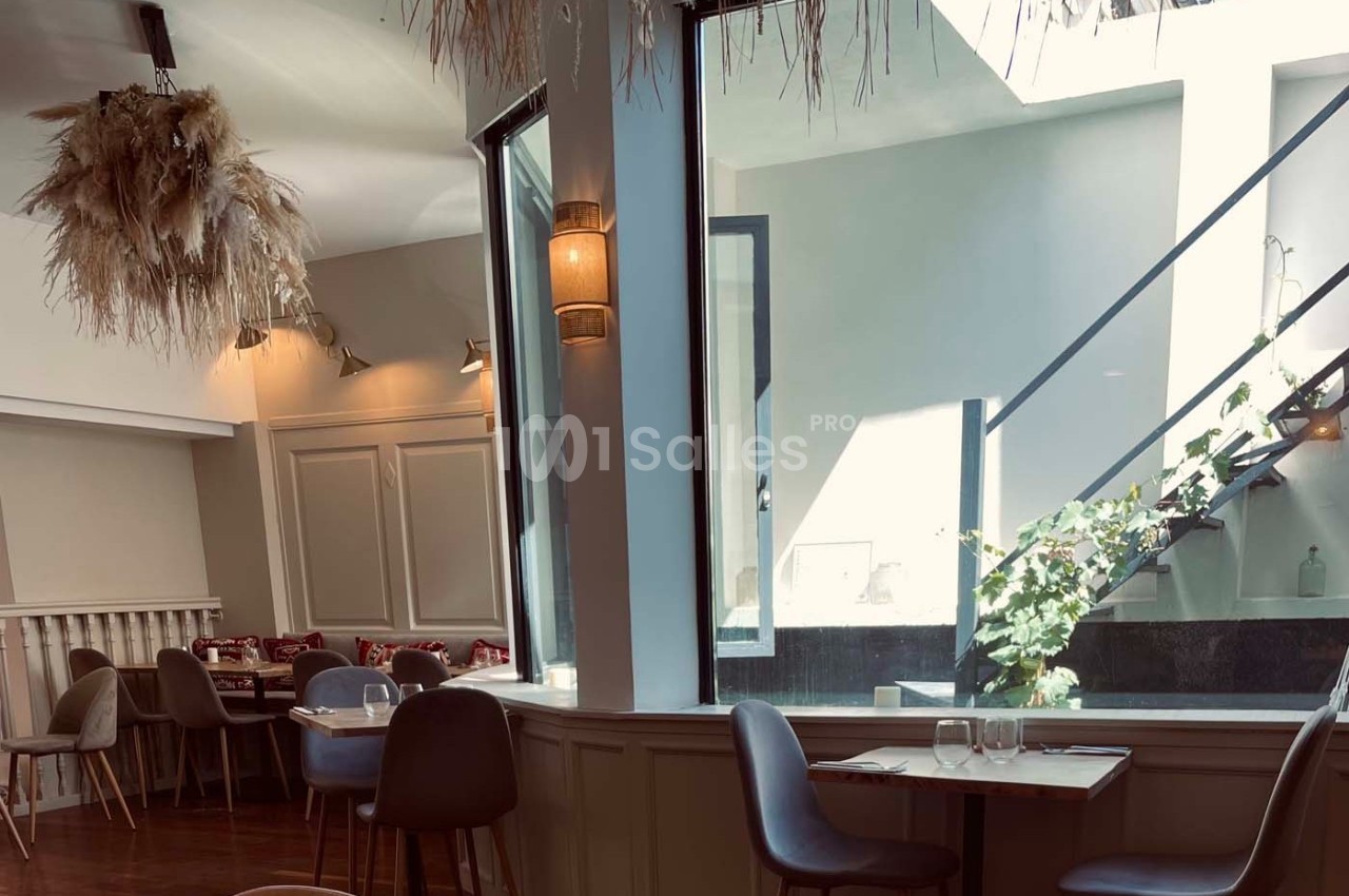 Salle de restaurant lumineuse avec des tables dressées, des chaises modernes et un escalier près d'une verrière.