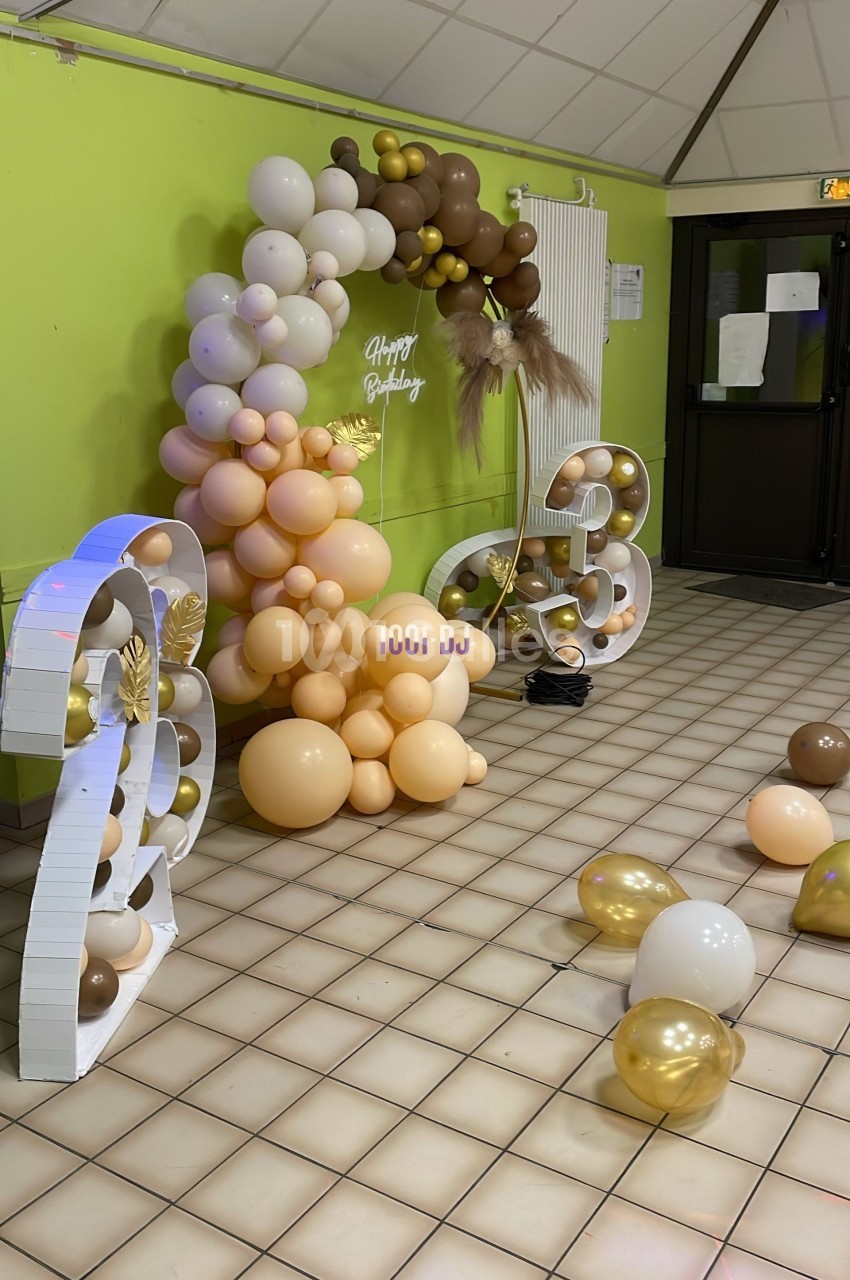 Décoration d'anniversaire avec arche de ballons beige et marron, chiffres lumineux et ballons dorés au sol.