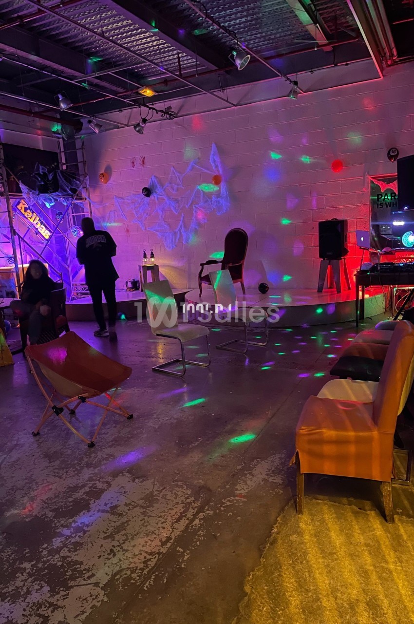 Salle industrielle avec éclairage coloré, chaises disposées, décorations et ambiance festive.