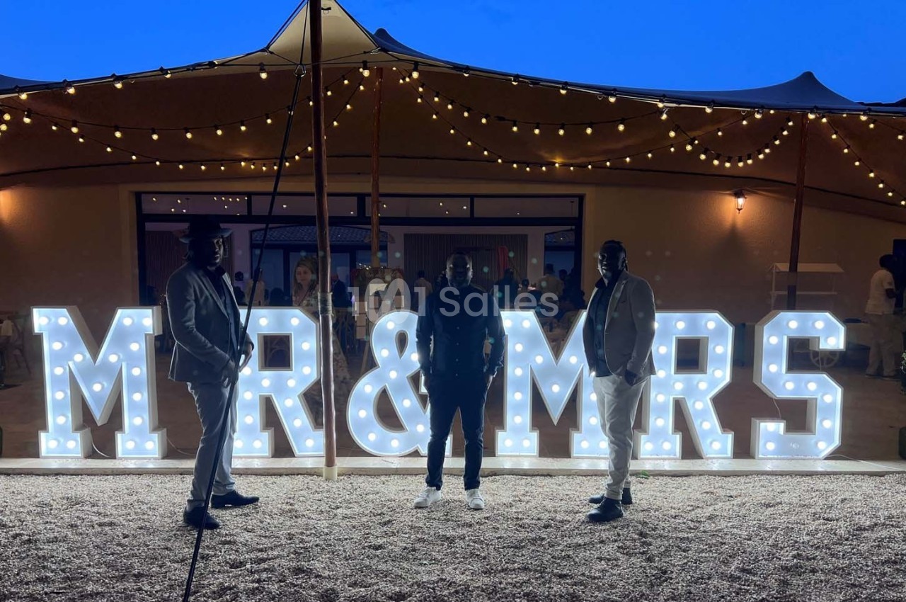 Trois personnes posent devant une enseigne lumineuse ’MR & MRS’ sous un ciel crépusculaire avec des guirlandes suspendues.