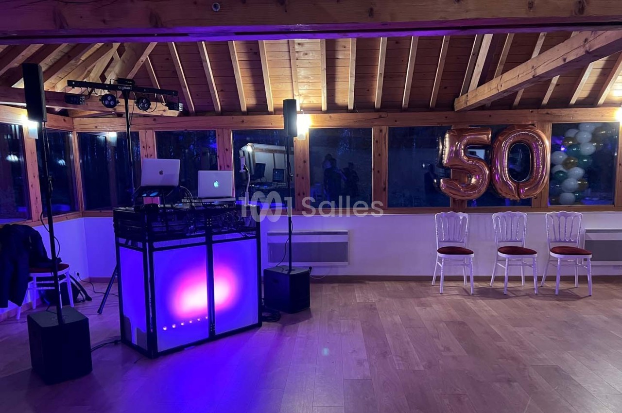 Salle décorée pour une fête, avec une table de DJ éclairée, ballons ’50’ et chaises alignées près des fenêtres.
