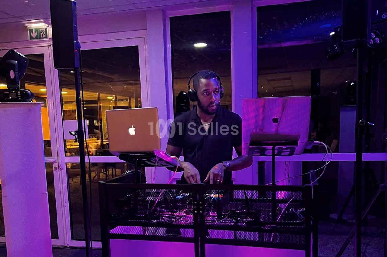 Un DJ mixe de la musique sur une table équipée d'ordinateurs et de matériel audio dans une salle éclairée en violet.