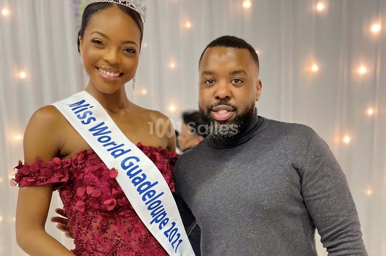 Une femme portant une écharpe ’Miss World Guadeloupe 2021’ pose avec un homme devant un fond lumineux.