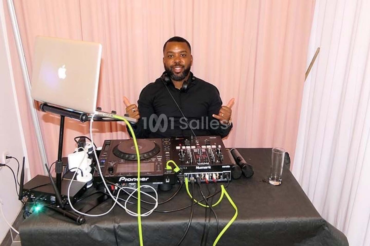 Un DJ souriant derrière une table équipée de matériel audio, avec un ordinateur portable et une table de mixage.