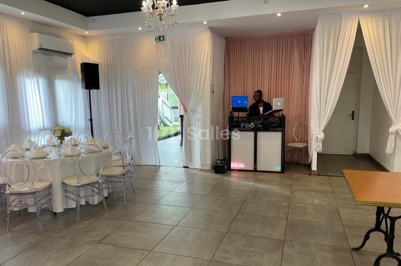 Salle de réception lumineuse avec table dressée, rideaux blancs et DJ installé près d'un mur rose.