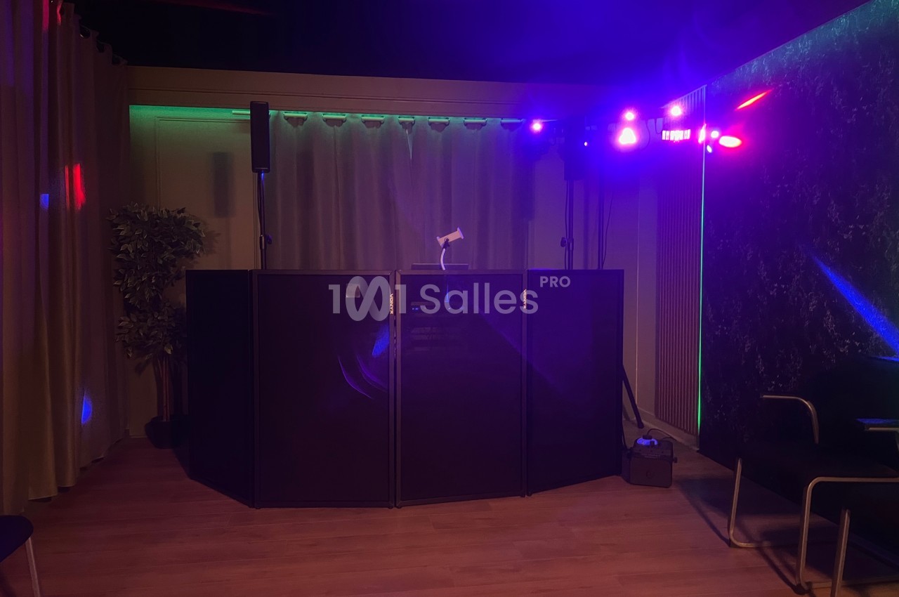 Console de DJ éclairée par des lumières colorées dans une salle sombre avec rideaux et plantes en arrière-plan.