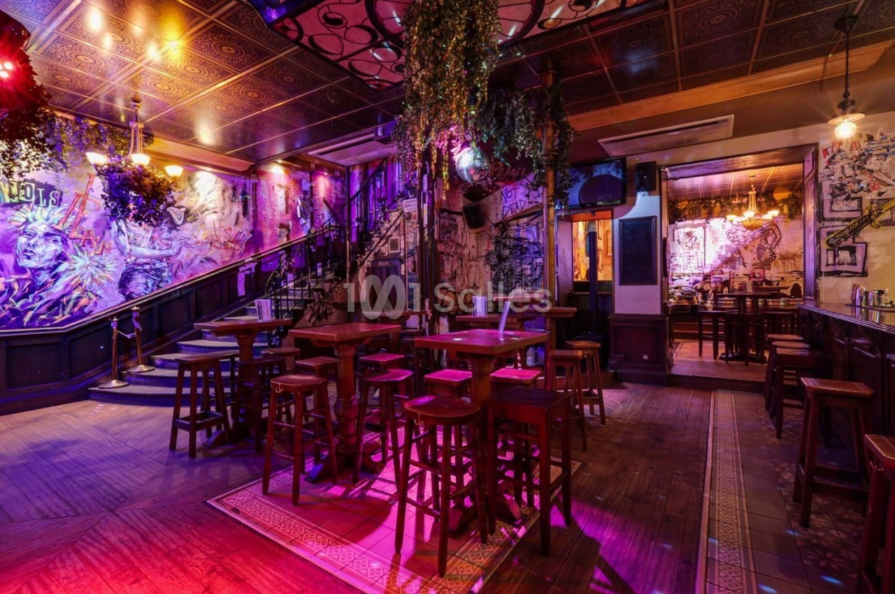 Intérieur d'un bar avec des tables hautes, des tabourets en bois, des lumières colorées et une décoration artistique.