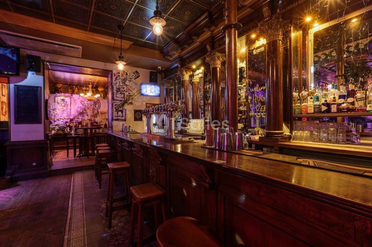 Intérieur d'un bar chaleureux avec comptoir en bois, tabourets, étagères de bouteilles et éclairage tamisé.