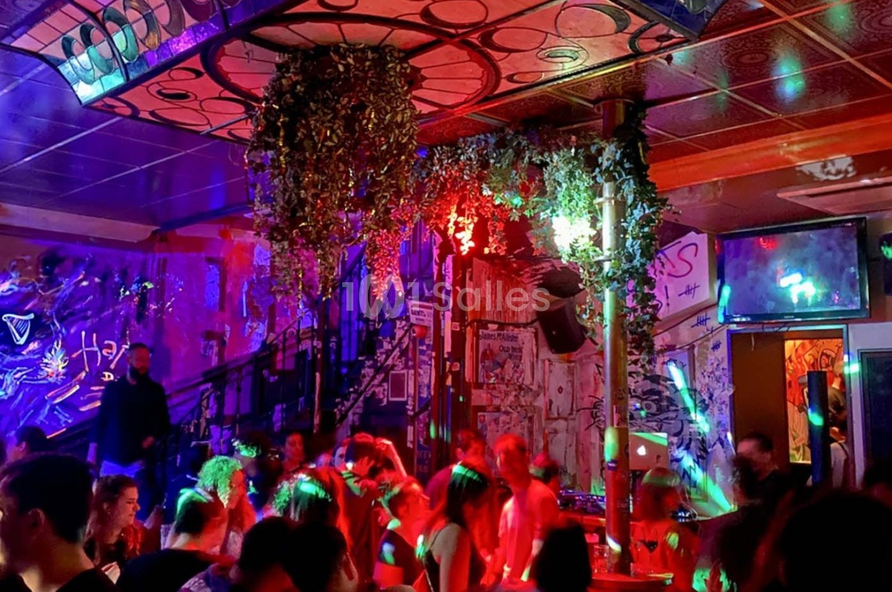 Salle animée avec des personnes dansant sous des lumières colorées, décorée de graffitis et de plantes suspendues.