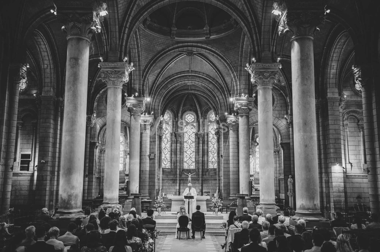 Intérieur d'une église avec des colonnes et des vitraux, une cérémonie en cours devant des personnes assises.
