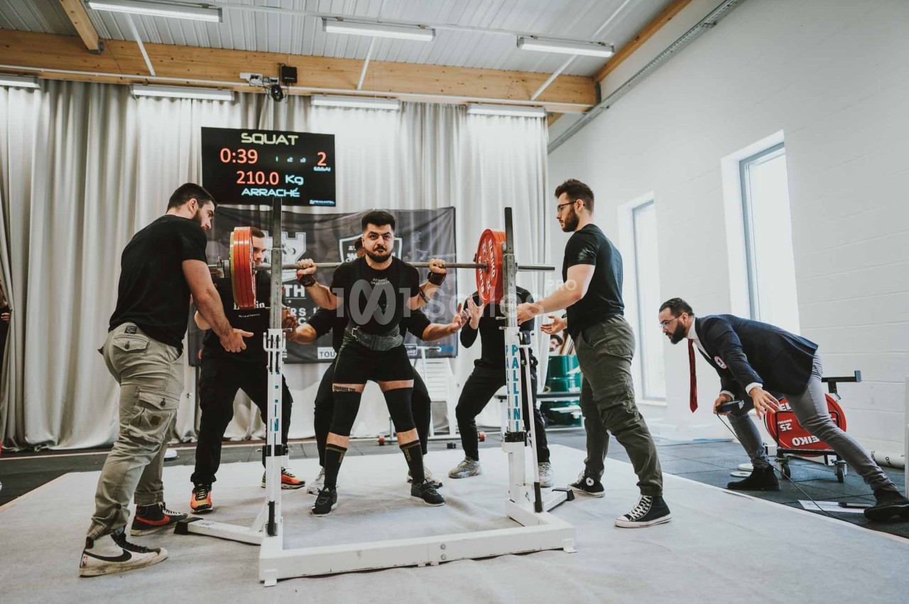Un homme réalise un squat avec une barre chargée, entouré de plusieurs assistants dans une salle de sport.