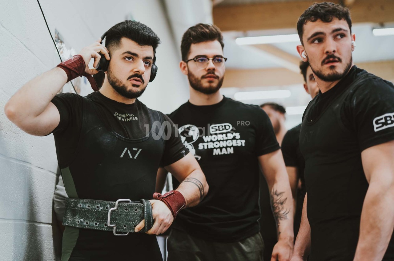 Trois hommes en tenue sportive, l'un ajustant sa ceinture de musculation, dans un environnement intérieur lumineux.