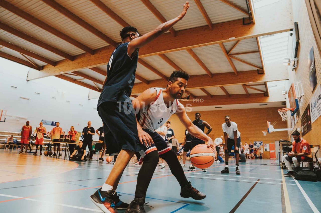 Deux joueurs de basketball s'affrontent sur un terrain intérieur, l'un dribblant tandis que l'autre défend.