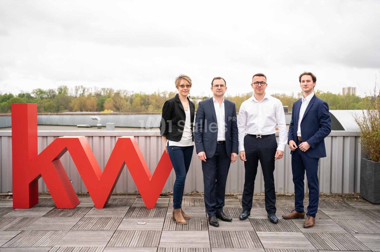 Quatre personnes posent debout sur une terrasse en bois près d'un grand logo rouge ’kw’.