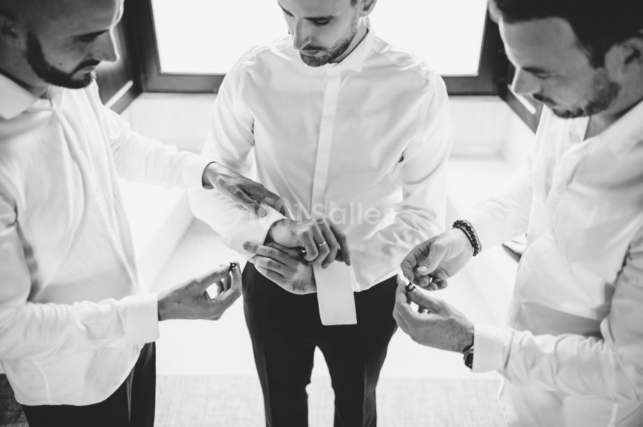 Trois hommes ajustent les accessoires et les manches de la chemise d'un quatrième homme dans une pièce lumineuse.