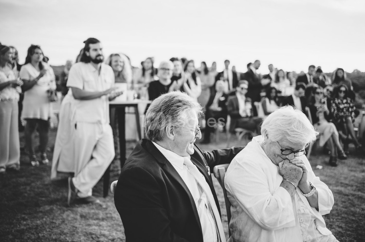Un couple âgé assis, échangeant un moment d'émotion, entouré d'invités lors d'un événement en plein air.