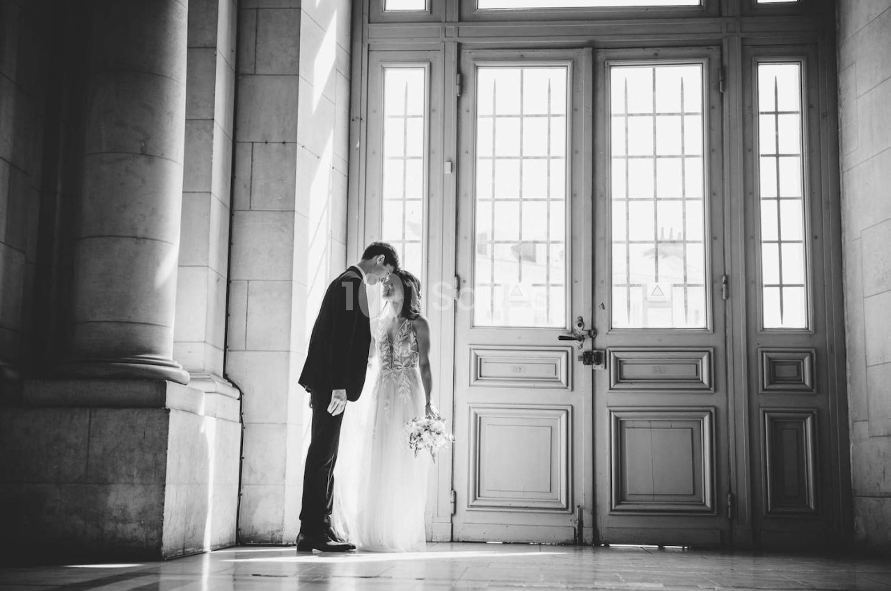 Un couple en tenue de mariage s'embrasse devant une grande porte vitrée dans un bâtiment lumineux.
