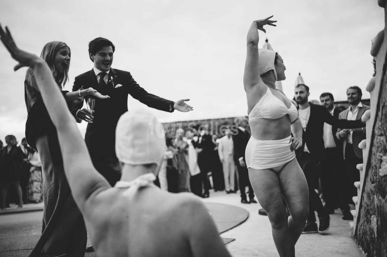 Des personnes en tenue élégante regardent des danseuses en maillot de bain et bonnet, lors d'un événement en extérieur.