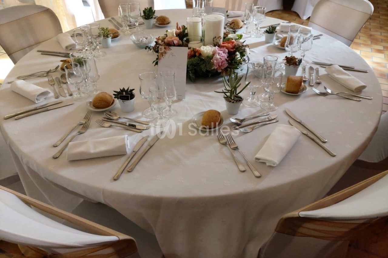Table ronde dressée pour un repas, avec nappes blanches, couverts, verres, bougies et centre de table floral.