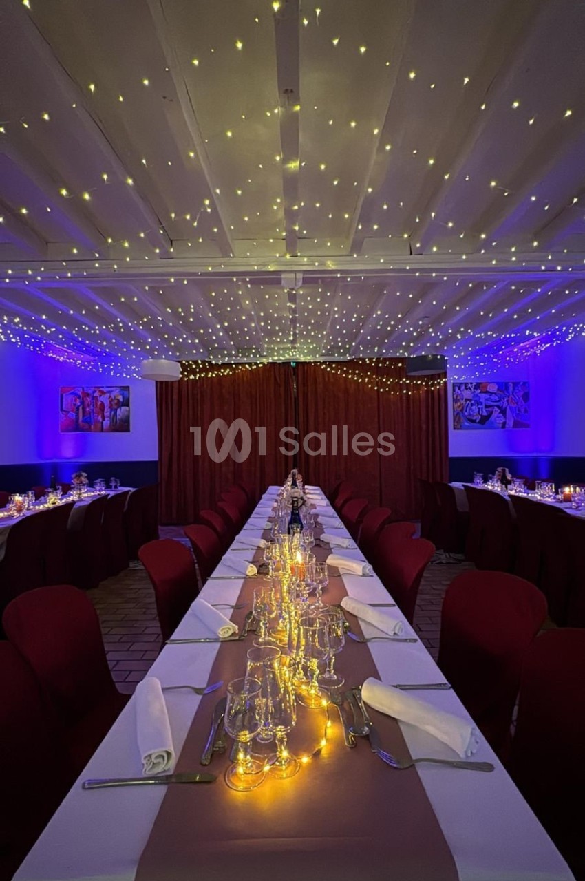 Salle de réception décorée avec une longue table dressée, nappes blanches, chaises rouges et guirlandes lumineuses au…