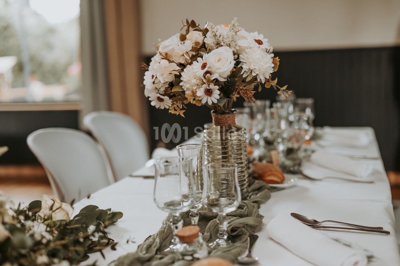 Centre de table floral avec des fleurs blanches et feuillage, disposé sur une table dressée avec vaisselle et serviettes.