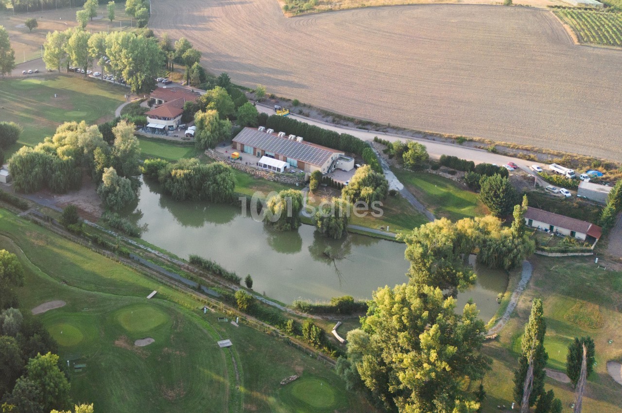 Vue aérienne d'un étang entouré de verdure, avec des bâtiments et des champs en arrière-plan.