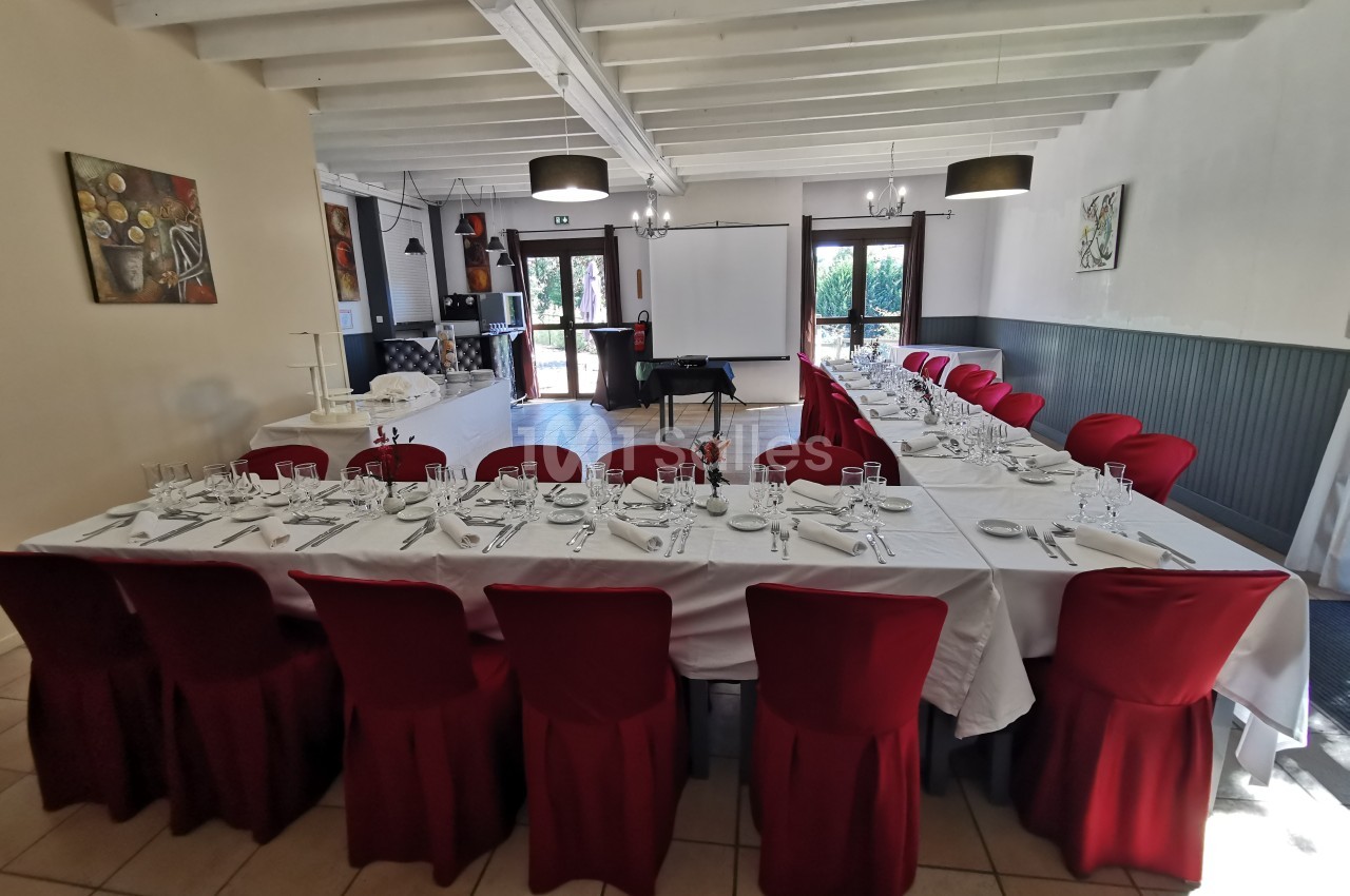 Salle de réception avec deux longues tables dressées, nappes blanches, chaises rouges et lumière naturelle.