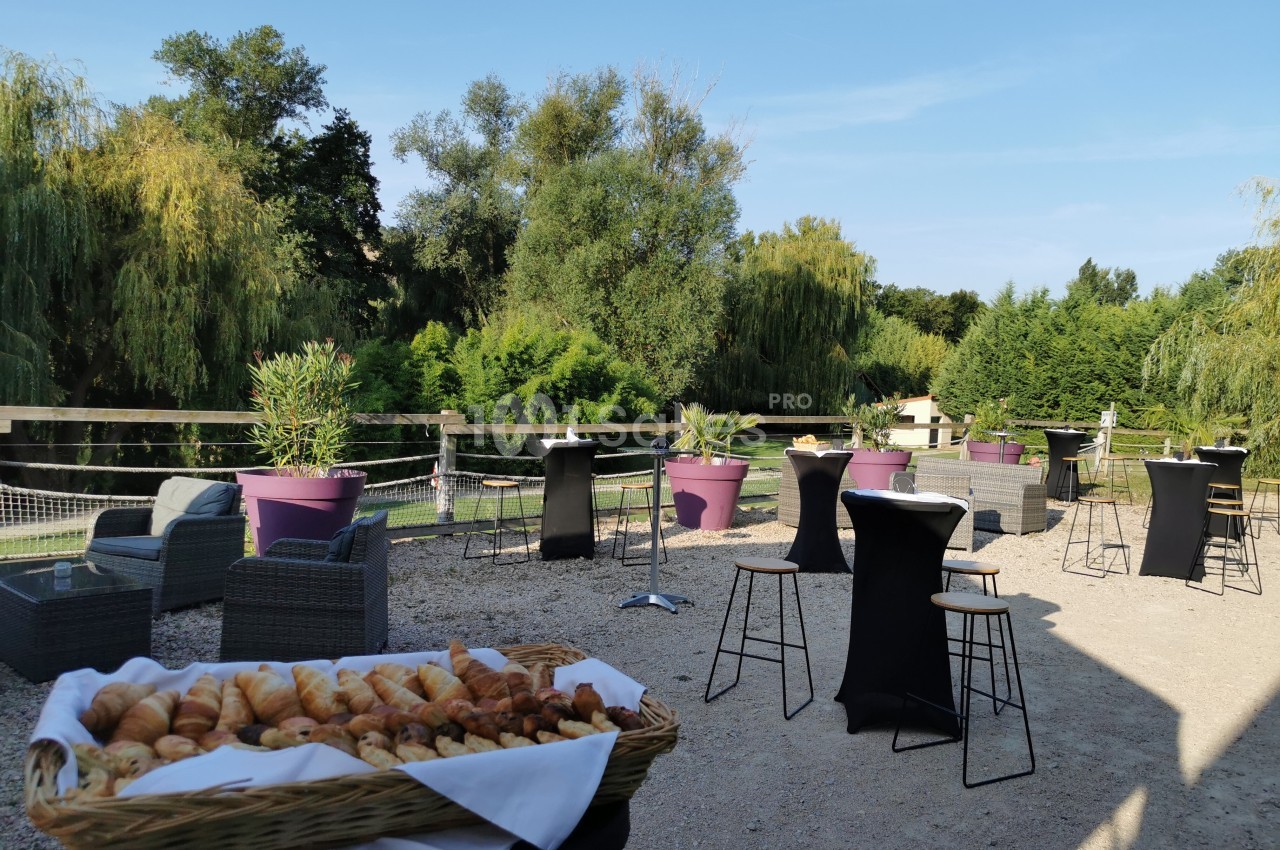 Terrasse extérieure avec tables hautes, fauteuils et un panier de viennoiseries, entourée de verdure et d'arbres.