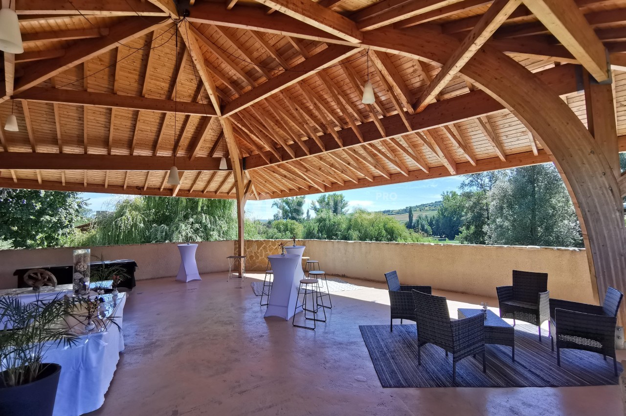 Terrasse couverte en bois avec vue sur un paysage verdoyant, aménagée avec tables hautes et fauteuils en rotin.
