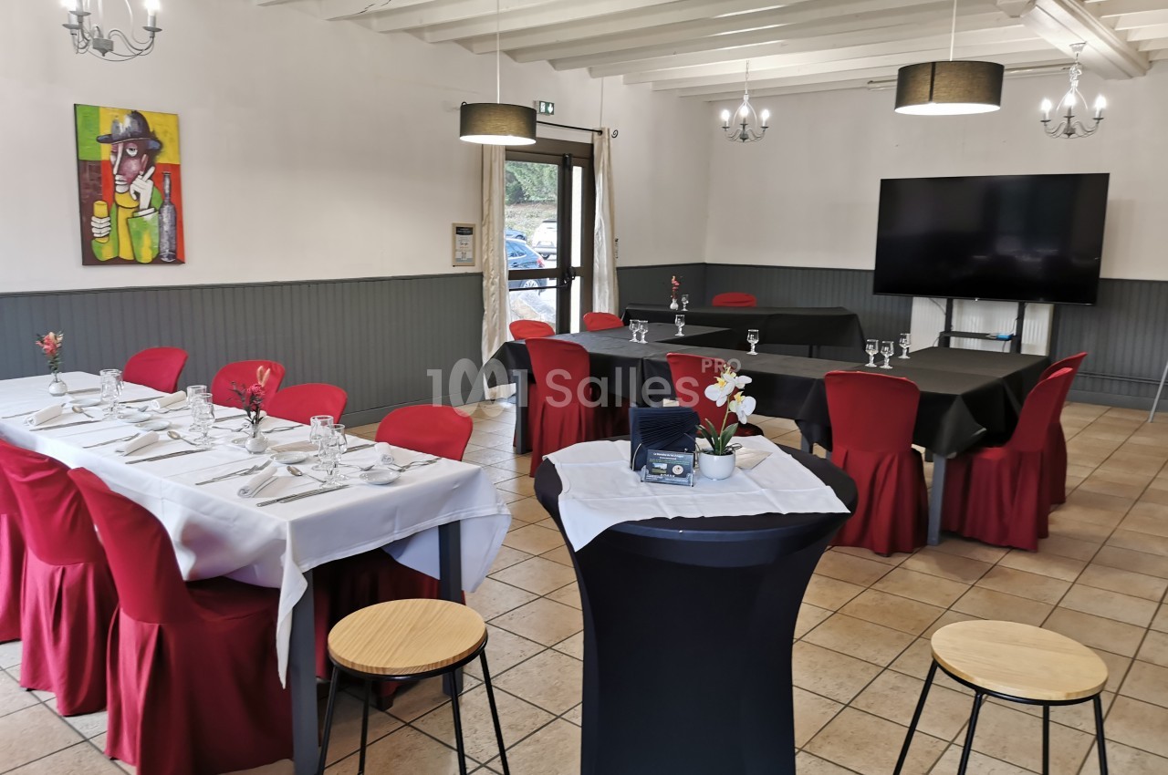 Salle lumineuse aménagée pour un événement, avec tables dressées, chaises rouges et décoration sobre.