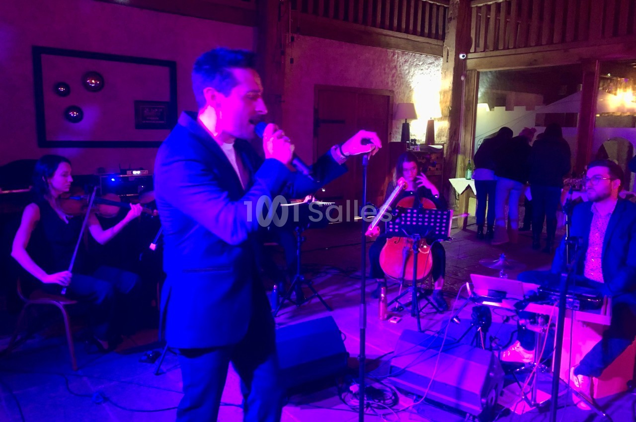 Un chanteur interprète sur scène accompagné de musiciens jouant du violon, violoncelle et clavier dans une salle éclairée.