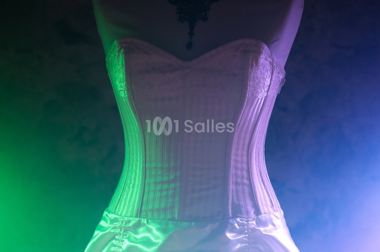 Corset blanc en dentelle et satin éclairé par des lumières vertes et violettes dans un environnement sombre.