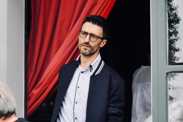 Homme portant des lunettes et une chemise claire, debout devant une porte avec un rideau rouge en arrière-plan.
