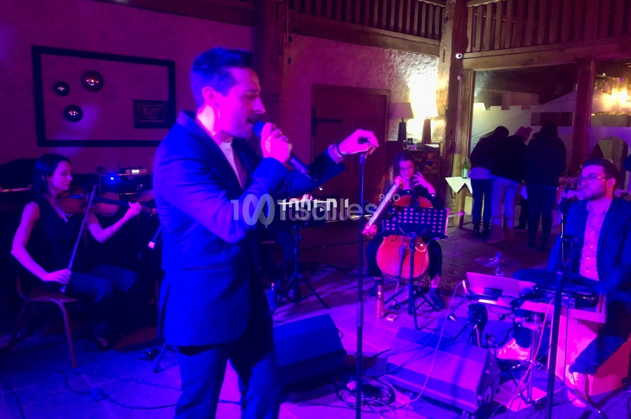 Un chanteur en costume interprète sur scène, accompagné de musiciens jouant du violon, violoncelle et clavier.