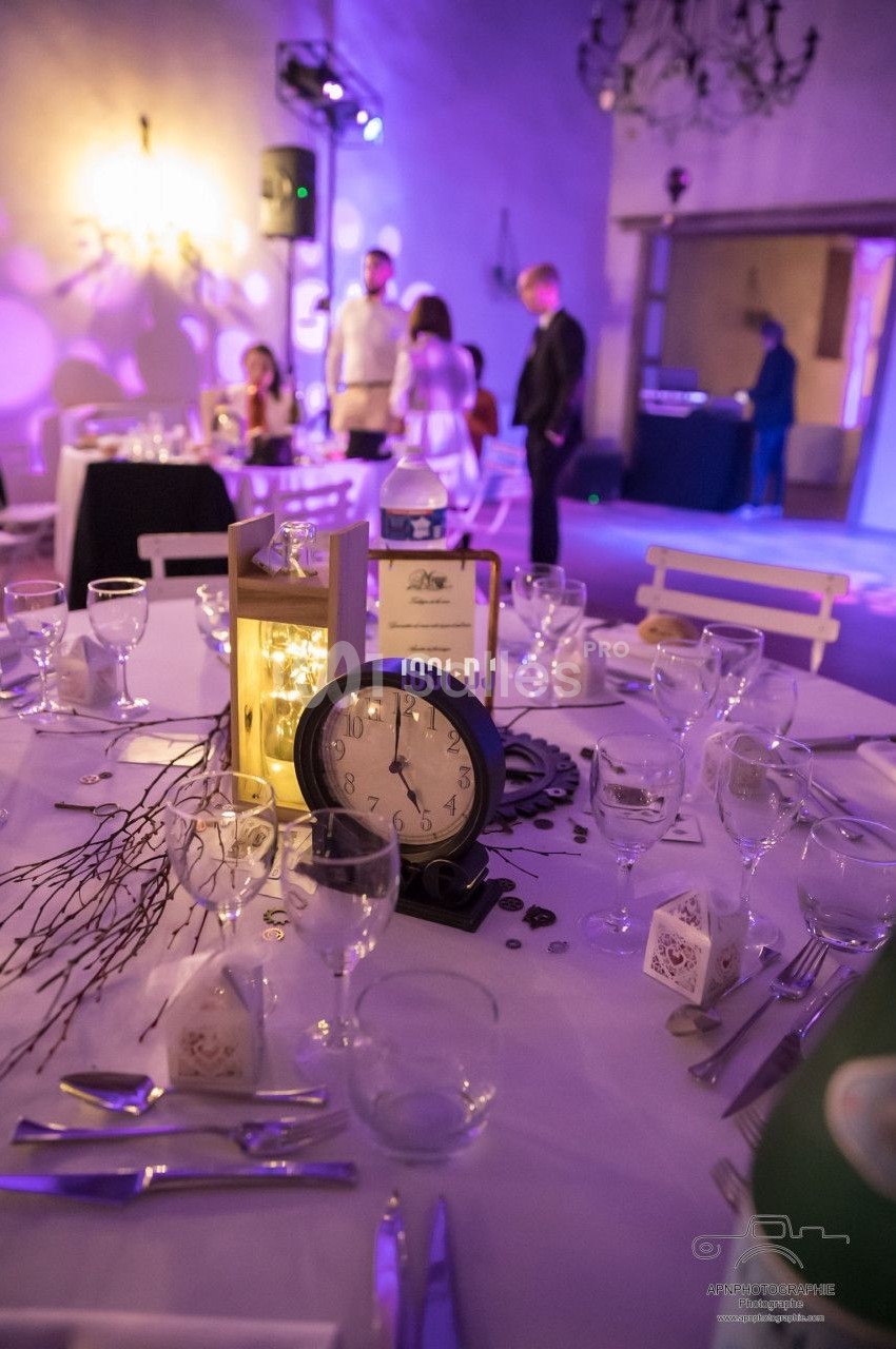 Table décorée pour un événement avec une horloge, des bougies et des branches, dans une salle éclairée en lumière tamisée.