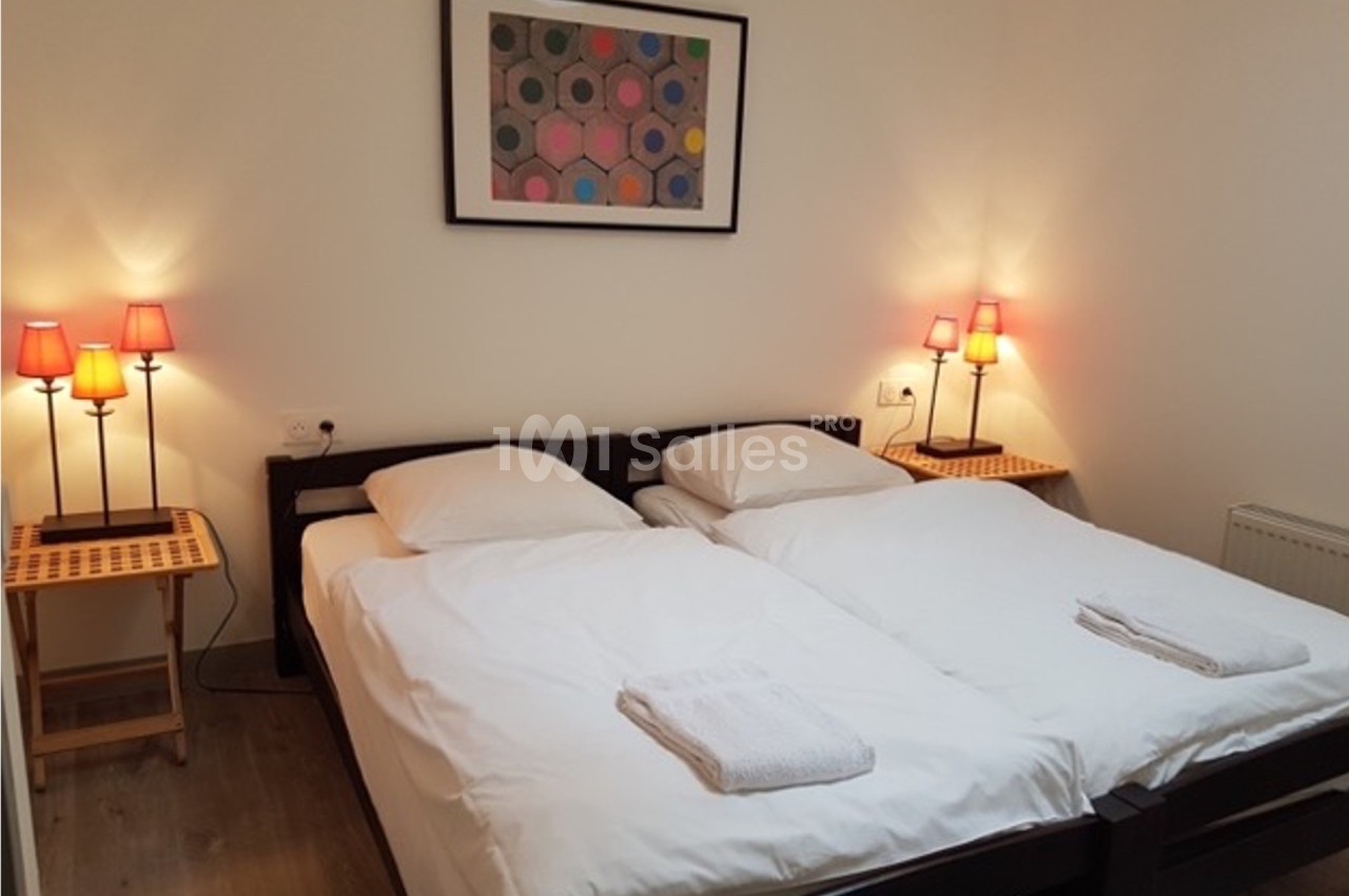 Deux lits simples avec draps blancs, tables de chevet en bois, lampes allumées et tableau coloré au mur.