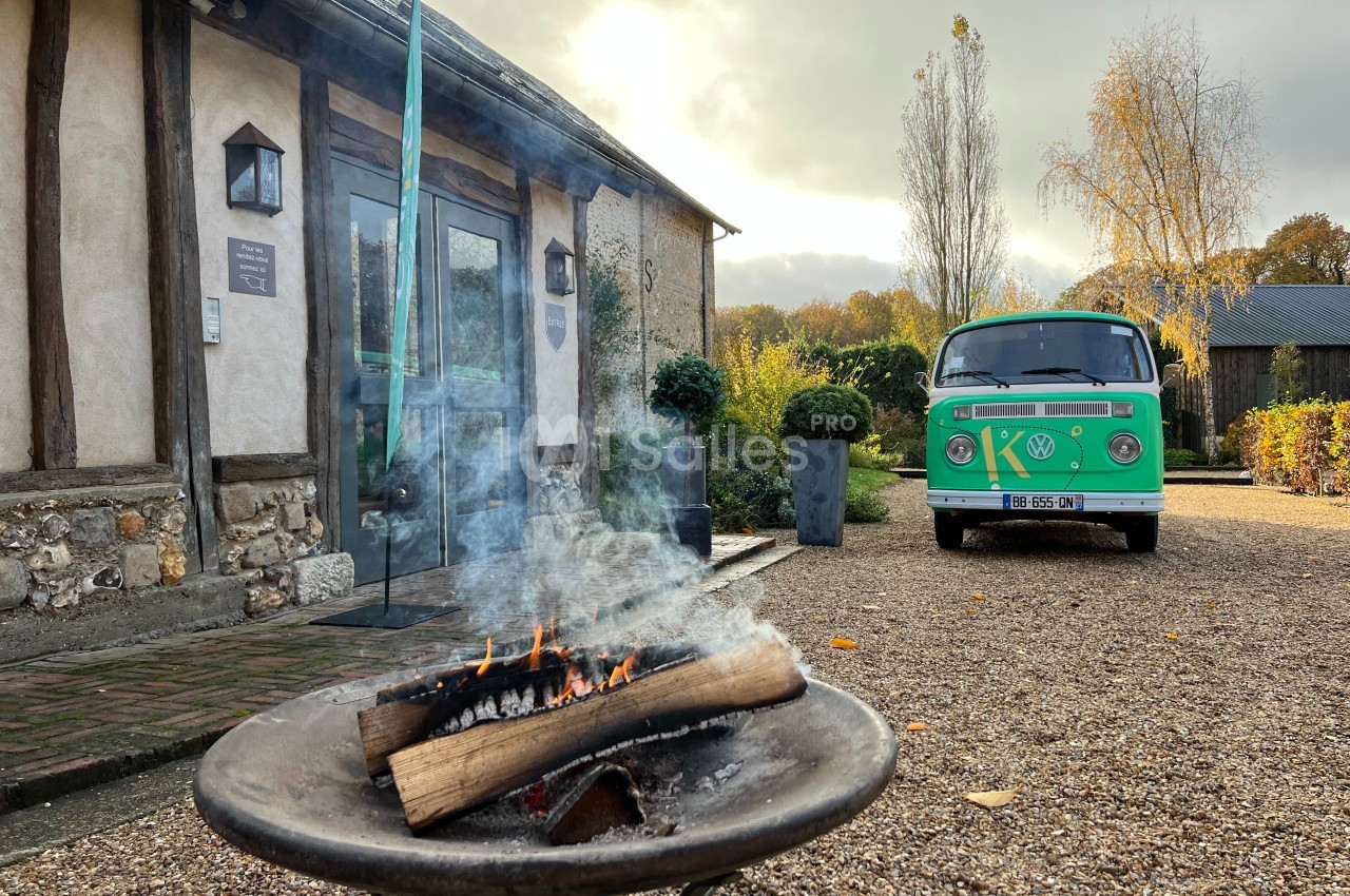 Brasero allumé devant une maison en pierre et bois, avec un van vert garé sur une allée gravillonnée.