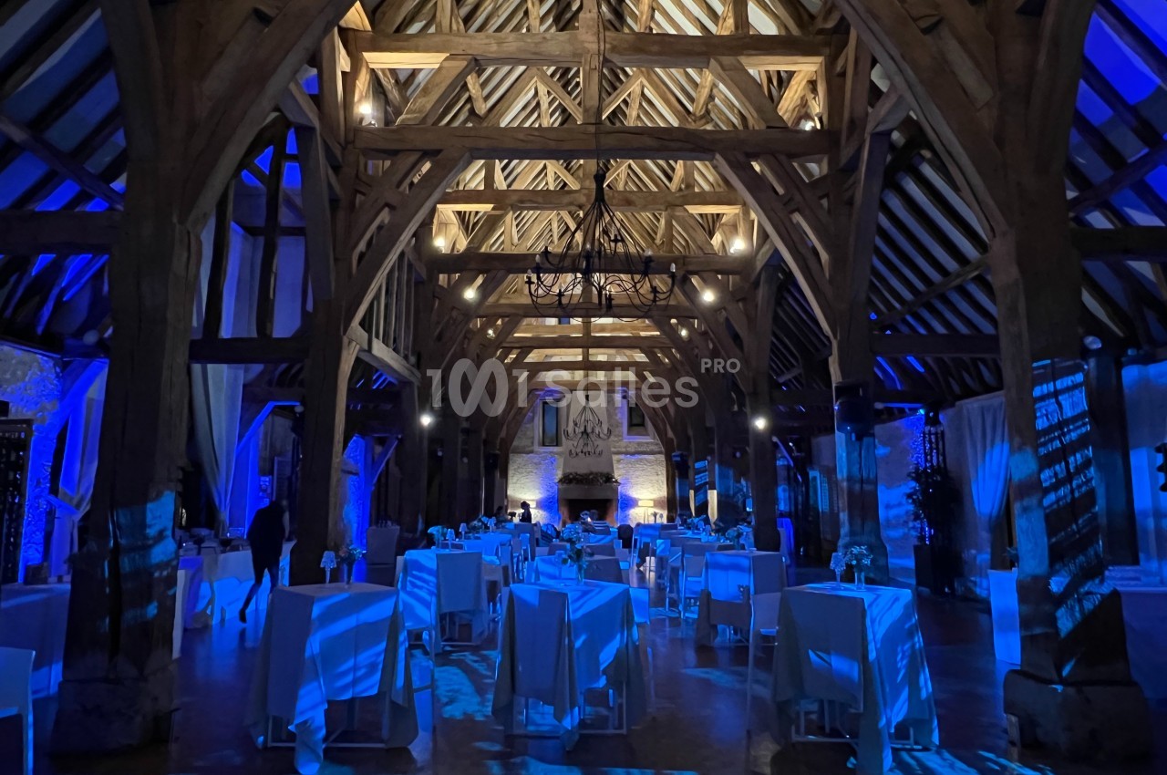 Salle de réception avec charpente en bois apparente, éclairage bleu et tables dressées pour un événement.
