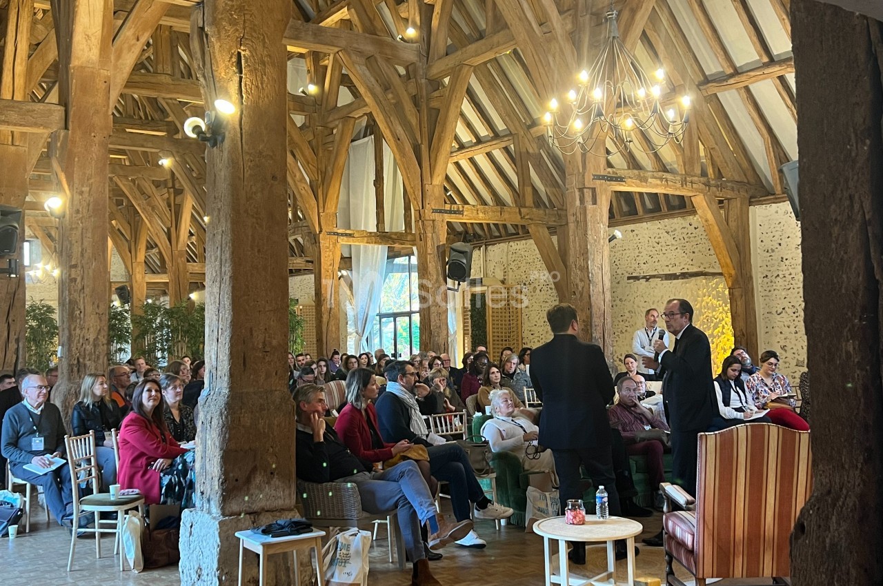 Un groupe de personnes assiste à une conférence dans une grande salle avec poutres apparentes et éclairage chaleureux.