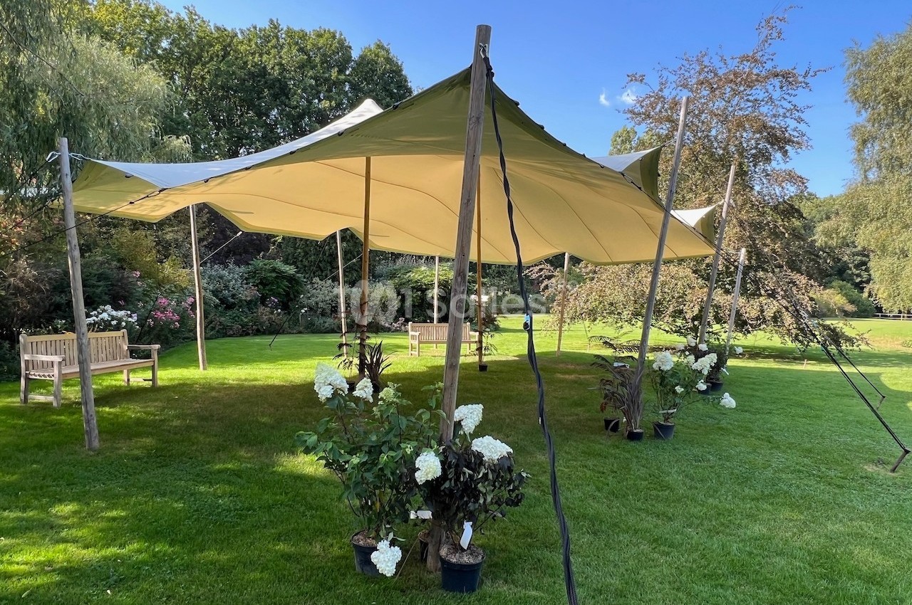 Tente beige montée sur une pelouse, entourée de plantes en pot et de bancs, dans un jardin arboré.