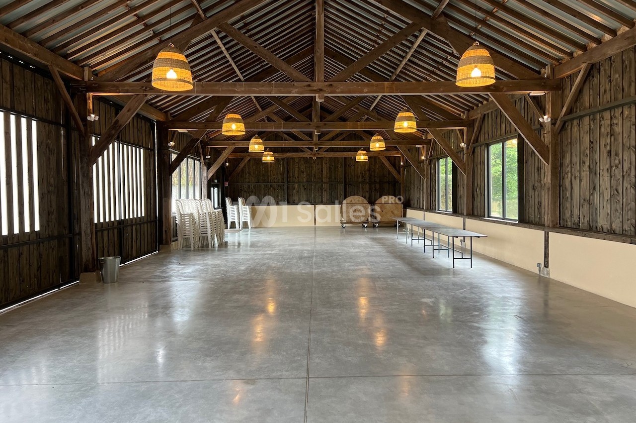 Intérieur d'une grande salle en bois avec plafond à poutres apparentes, sol en béton et suspensions lumineuses.