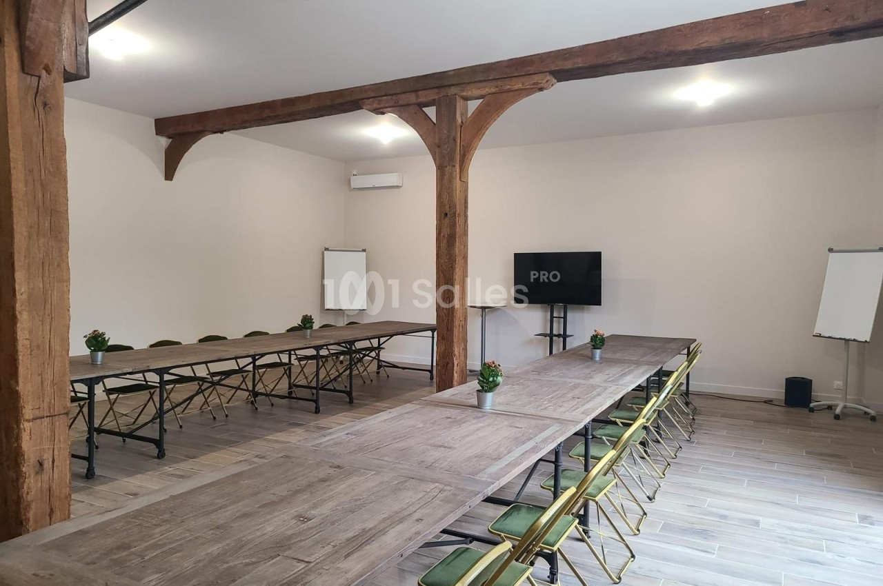 Salle de réunion lumineuse avec tables en bois disposées en U, chaises pliantes, écran, paperboards et poutres apparentes.