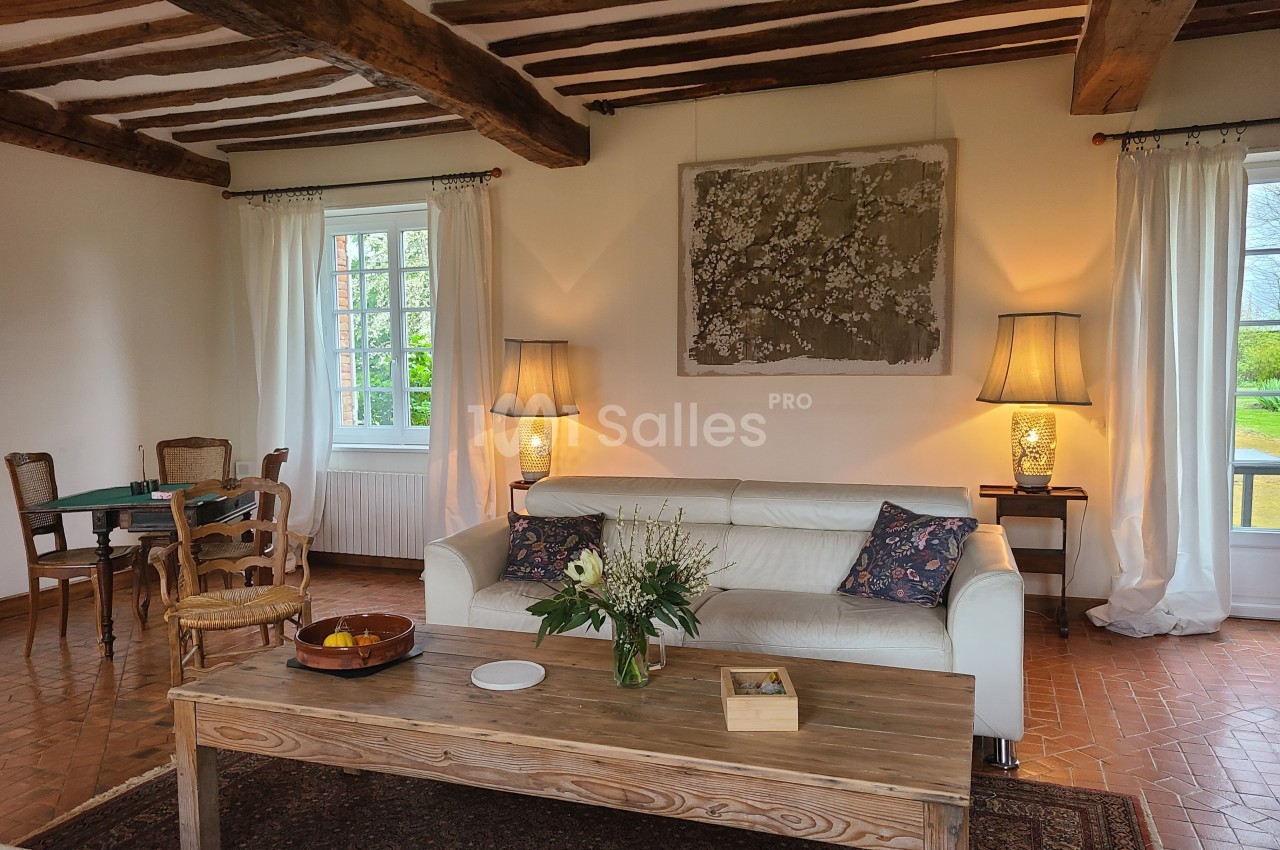 Salon lumineux avec poutres apparentes, canapé blanc, table basse en bois, lampes allumées et vue sur jardin.