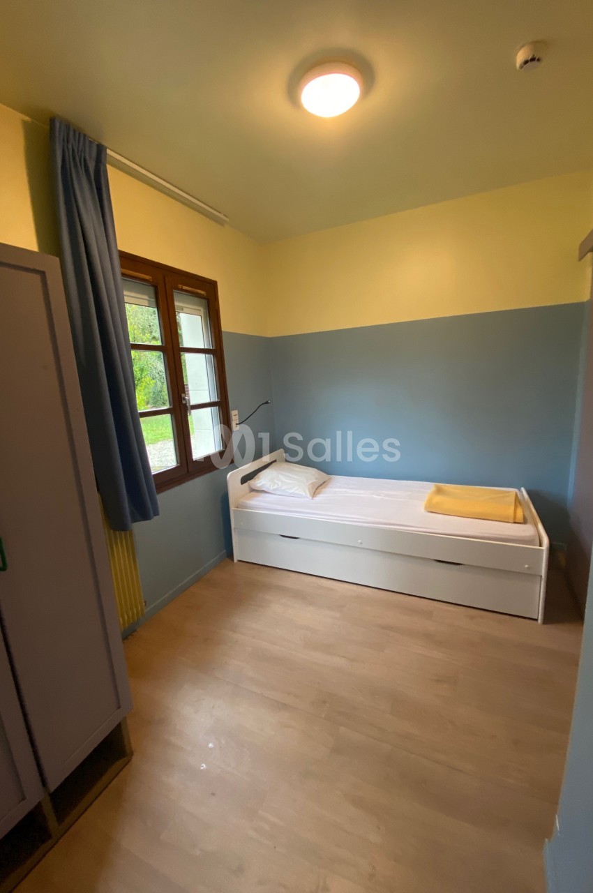 Chambre simple avec lit une place, armoire, fenêtre avec rideau bleu et murs peints en jaune et bleu.