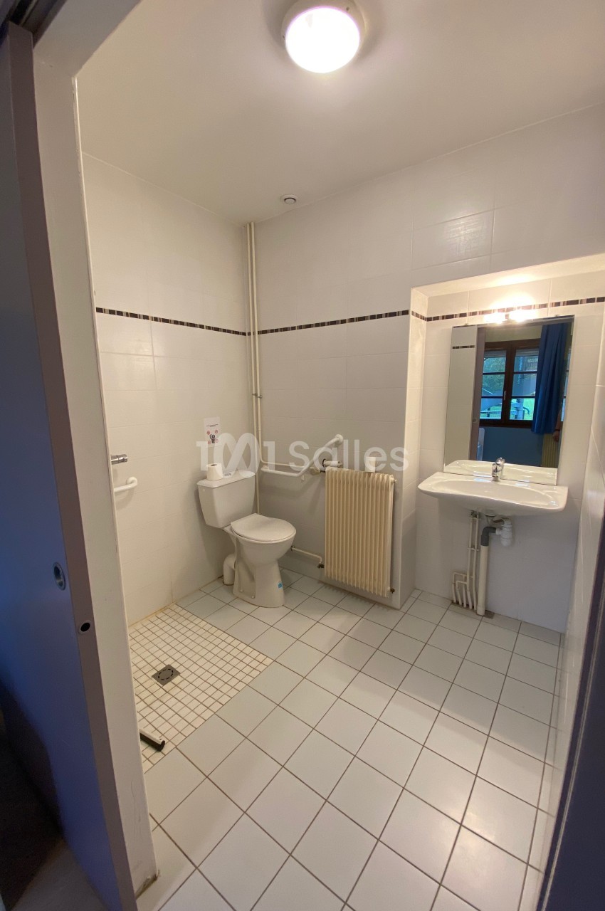 Salle de bain accessible avec toilettes, barre d'appui, lavabo, miroir et carrelage clair.