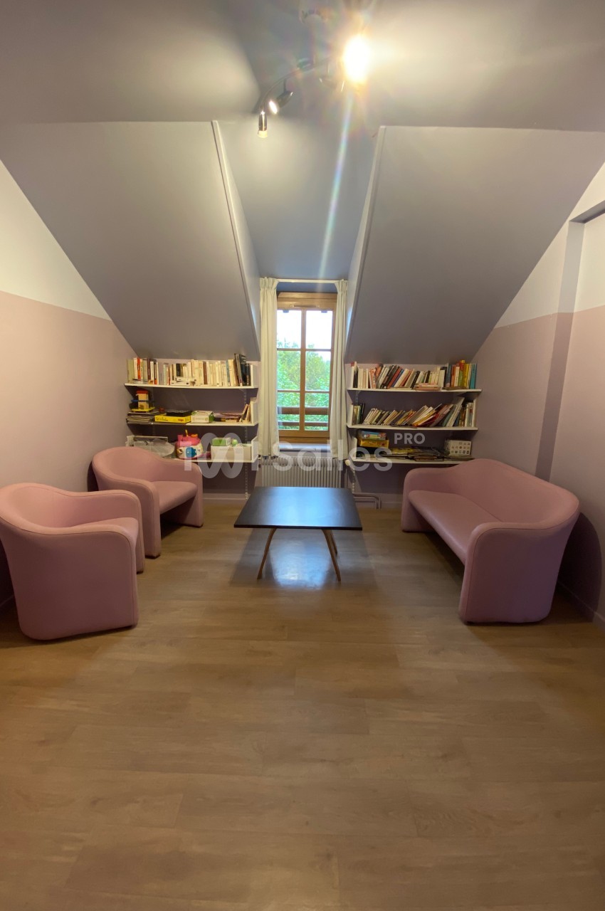 Salon mansardé avec murs violets, deux fauteuils, un canapé rose, une table basse et des étagères remplies de livres.