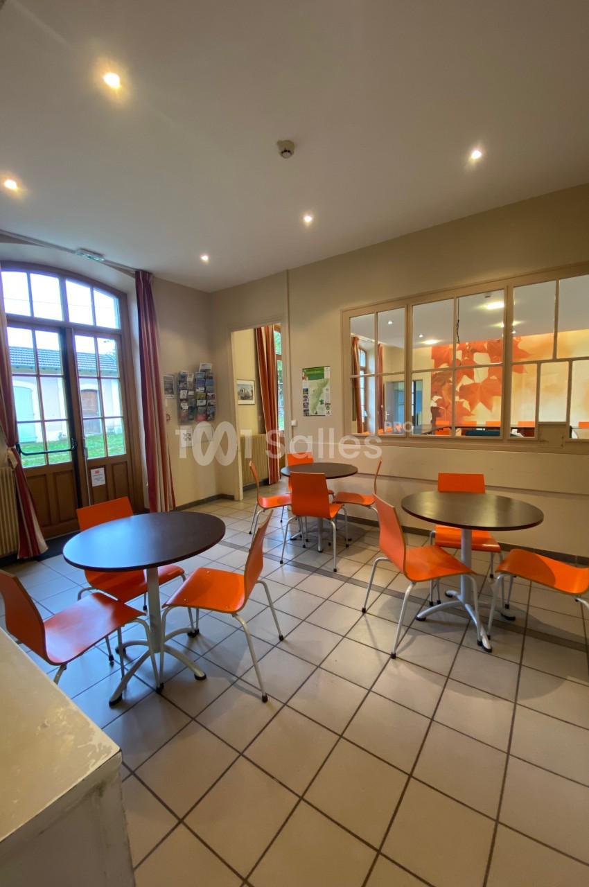 Salle lumineuse avec tables rondes, chaises orange, carrelage clair, grandes fenêtres et panneaux d'affichage au mur.