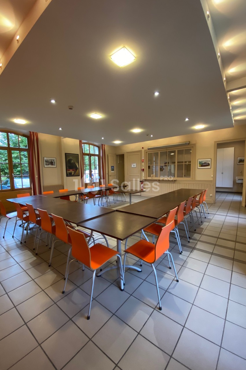 Salle de réunion lumineuse avec tables disposées en U et chaises orange, entourée de grandes fenêtres.